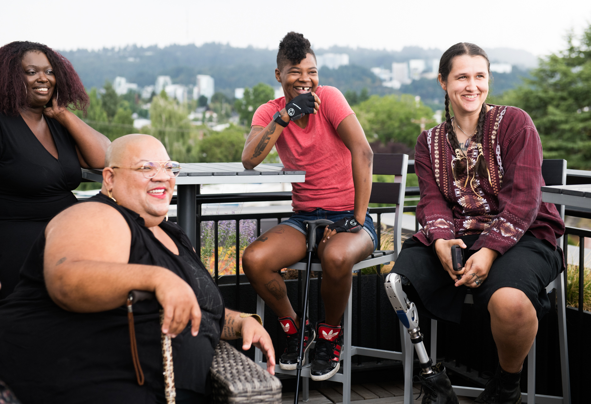 Four disabled people of color with canes, prosthetic legs, and a wheelchair sit on a rooftop deck, laughing and sharing stories. Greenery and city high-rises are visible in the background.