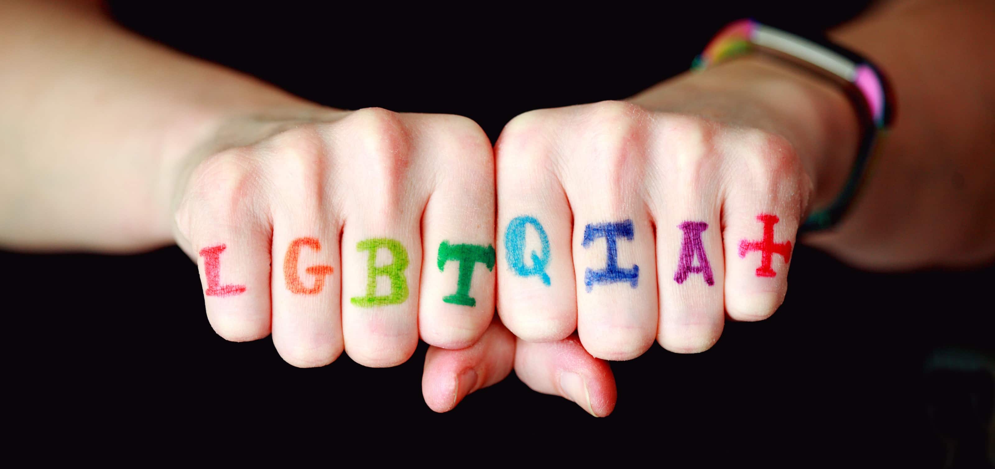 Photo of two fists together with the letters LGBTQIA+ on each finger in rainbow colors 